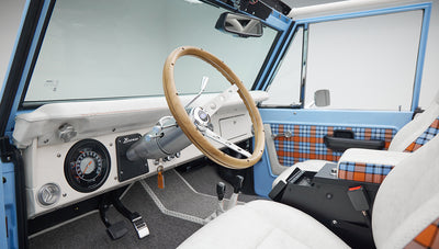 Interior of Classic Ford Bronco showing wooden steering wheel, white seats, gray flooring, and plaid door panel