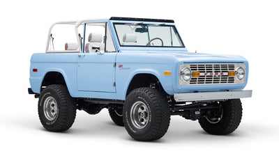 Classic Ford Bronco in light blue with white roll bar and plaid seat covers, front three-quarter view