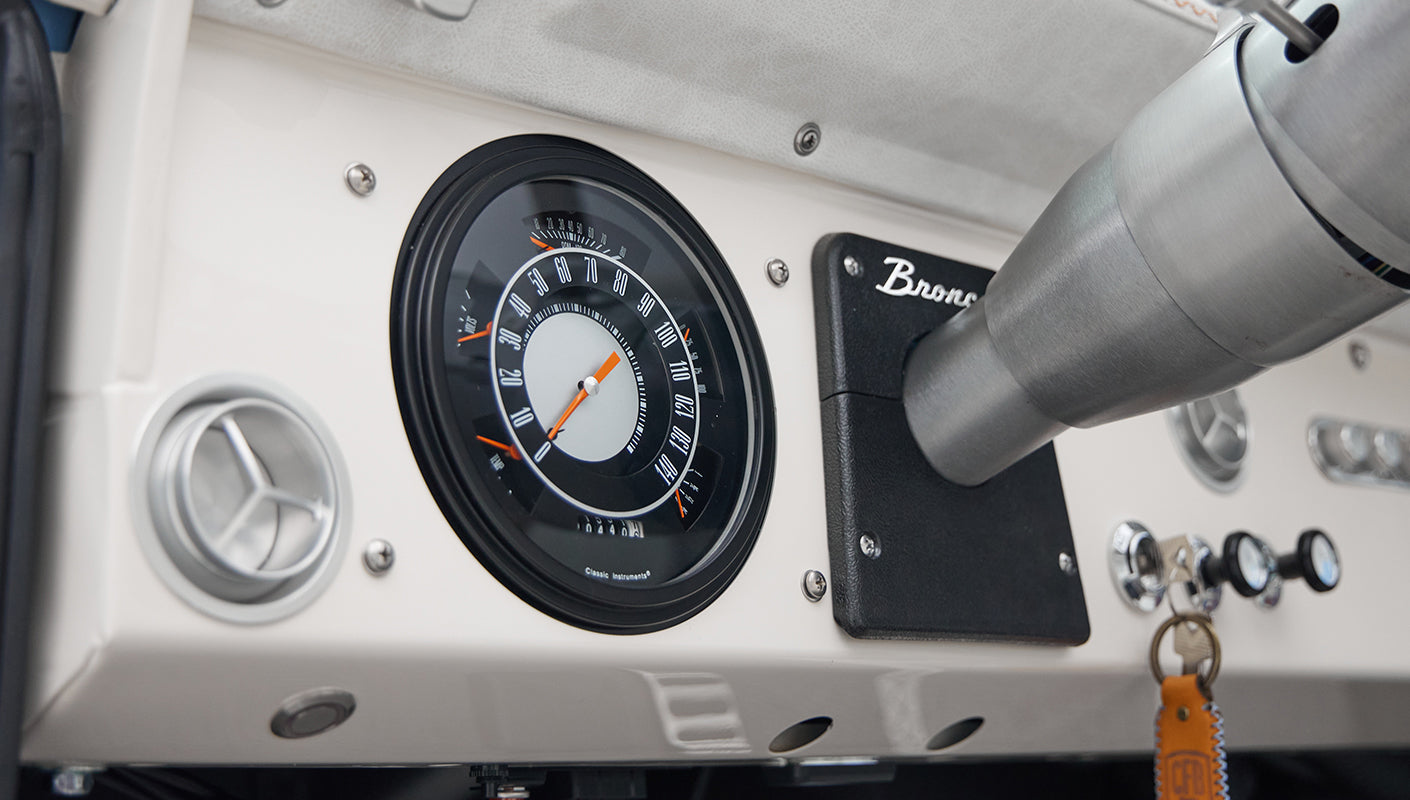 Round black speedometer gauge and Bronco plaque on white dashboard near steering column inside Classic Ford Bronco
