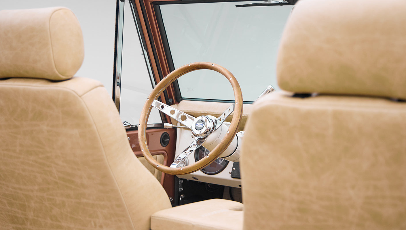 View from rear seat showing wood steering wheel and beige interior of Classic Ford Bronco