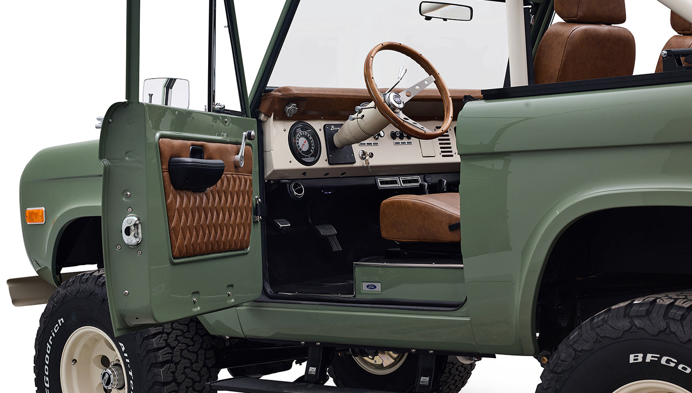 Open driver-side door showing interior of Classic Ford Bronco with brown leather seats and wood steering wheel