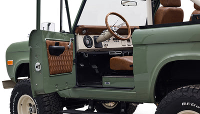 Open driver-side door showing interior of Classic Ford Bronco with brown leather seats and wood steering wheel