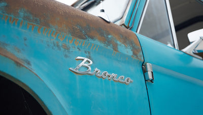 Close-up of rusted turquoise Classic Ford Bronco door with chrome "Bronco" emblem.