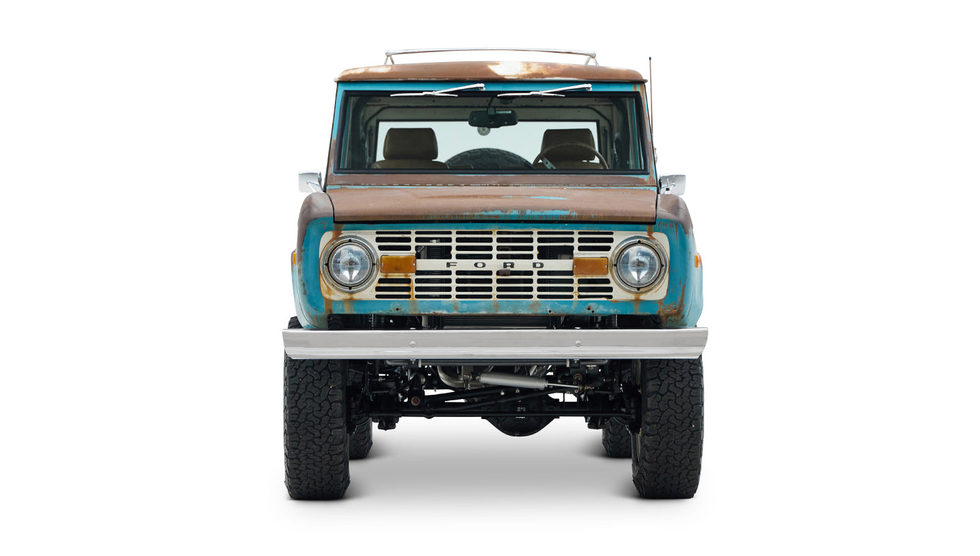 Frontal view of a rusted turquoise Classic Ford Bronco showing grill, headlights, and rugged tires.