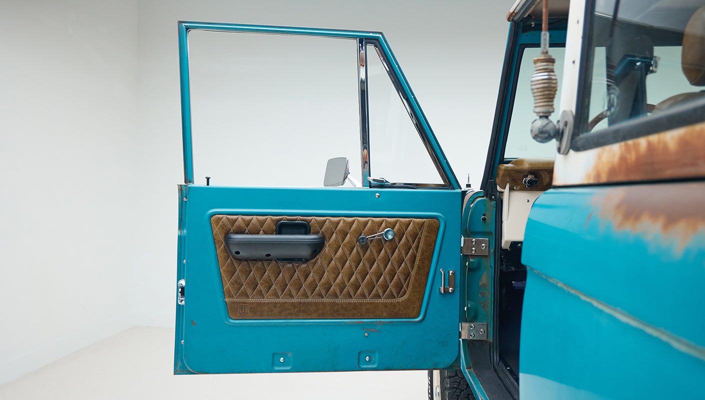 Open driver side door of Classic Ford Bronco showing turquoise exterior and brown diamond-stitched interior panel