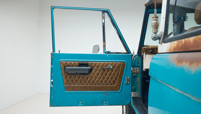 Open driver side door of Classic Ford Bronco showing turquoise exterior and brown diamond-stitched interior panel
