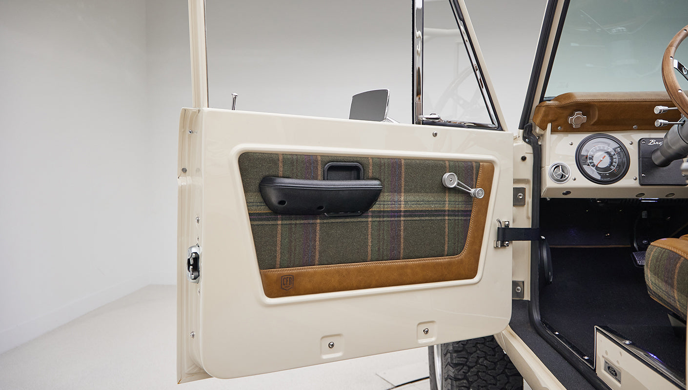 Open driver side door of Classic Ford Bronco with plaid and leather panel face and window crank