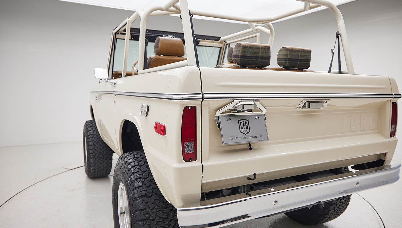 Rear-side view of cream-colored Classic Ford Bronco showing open roll cage and plaid headrests