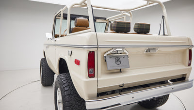 Rear-side view of cream-colored Classic Ford Bronco showing open roll cage and plaid headrests