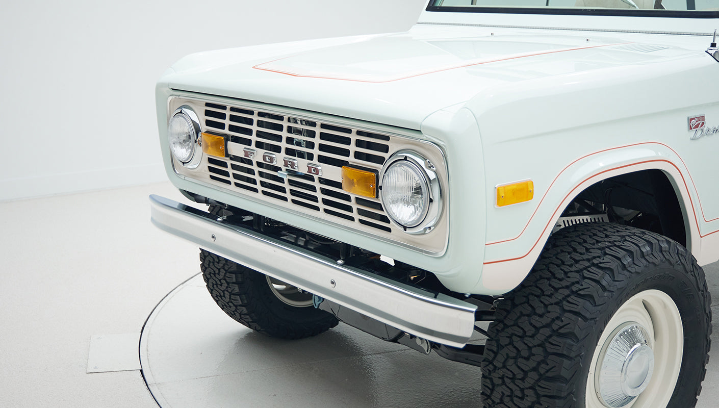 Front left side of a Classic Ford Bronco showing grill, headlights, and off-road tires on a white floor.