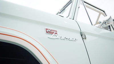 Close-up of Classic Ford Bronco emblem and part of the front fender with pale blue paint and red pinstriping.