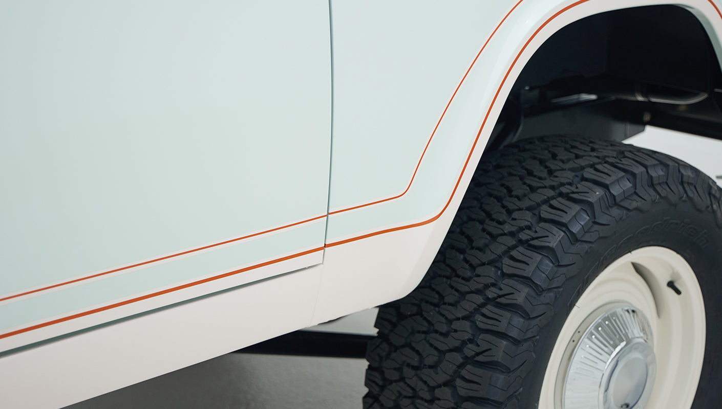 Lower side panel of Classic Ford Bronco showing pale blue and white paint separated by thin red stripes over an off-road