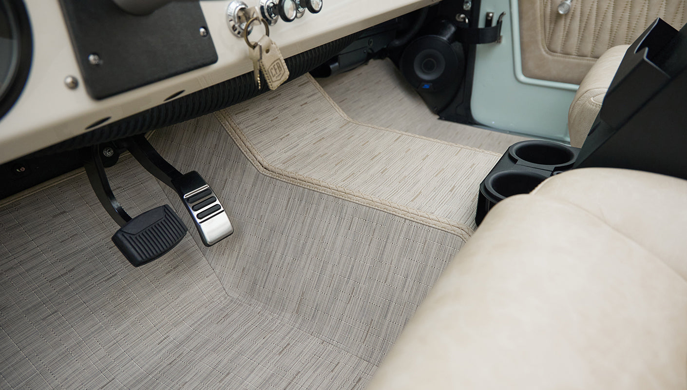Floor and pedal area of Classic Ford Bronco with patterned light beige carpet and beige seat side visible.