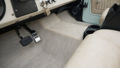 Floor and pedal area of Classic Ford Bronco with patterned light beige carpet and beige seat side visible.