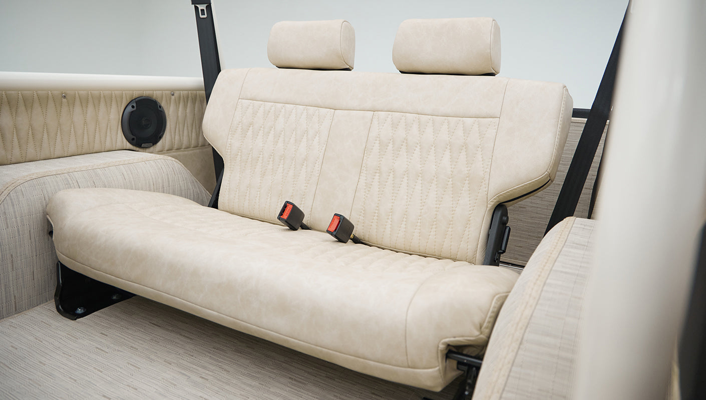 Rear interior bench seat in beige upholstery inside Classic Ford Bronco with seat belts and patterned flooring.