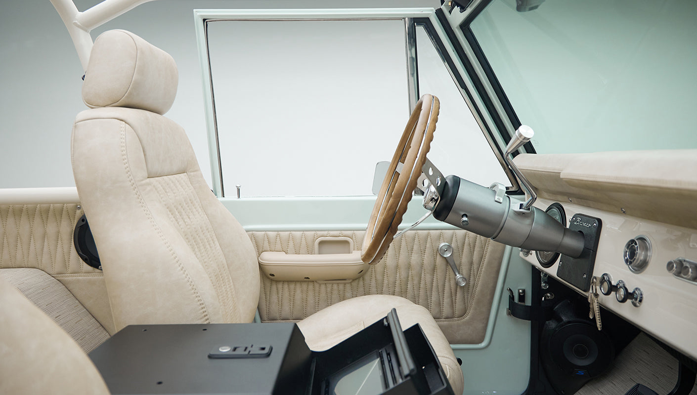Classic Ford Bronco driver side interior view showing beige seats, wood steering wheel, and speaker under dashboard.