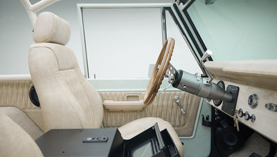Classic Ford Bronco driver side interior view showing beige seats, wood steering wheel, and speaker under dashboard.