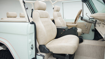 Side view of Classic Ford Bronco's front seats and interior in beige upholstery with white exterior and roll cage.