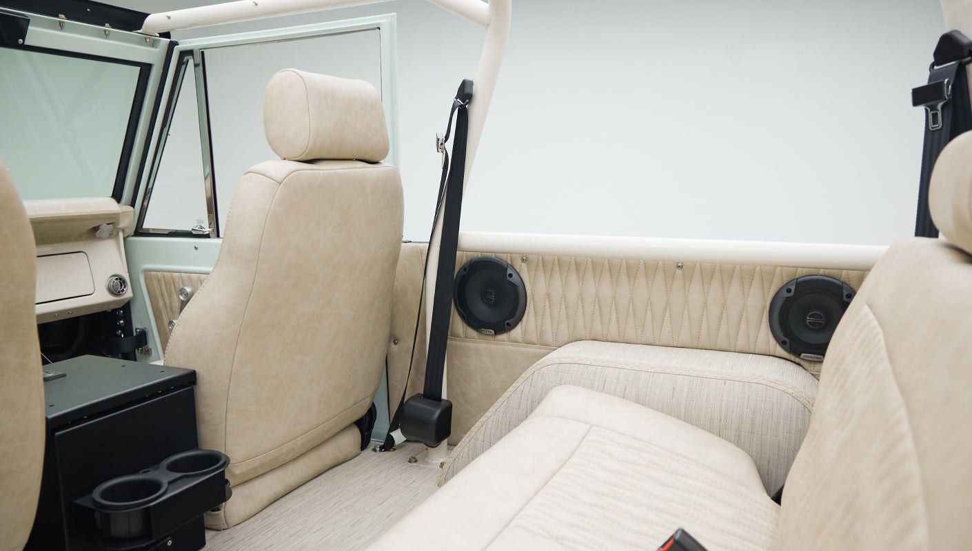 Interior of Classic Ford Bronco showing beige seats, speakers in side panels, and seat belts