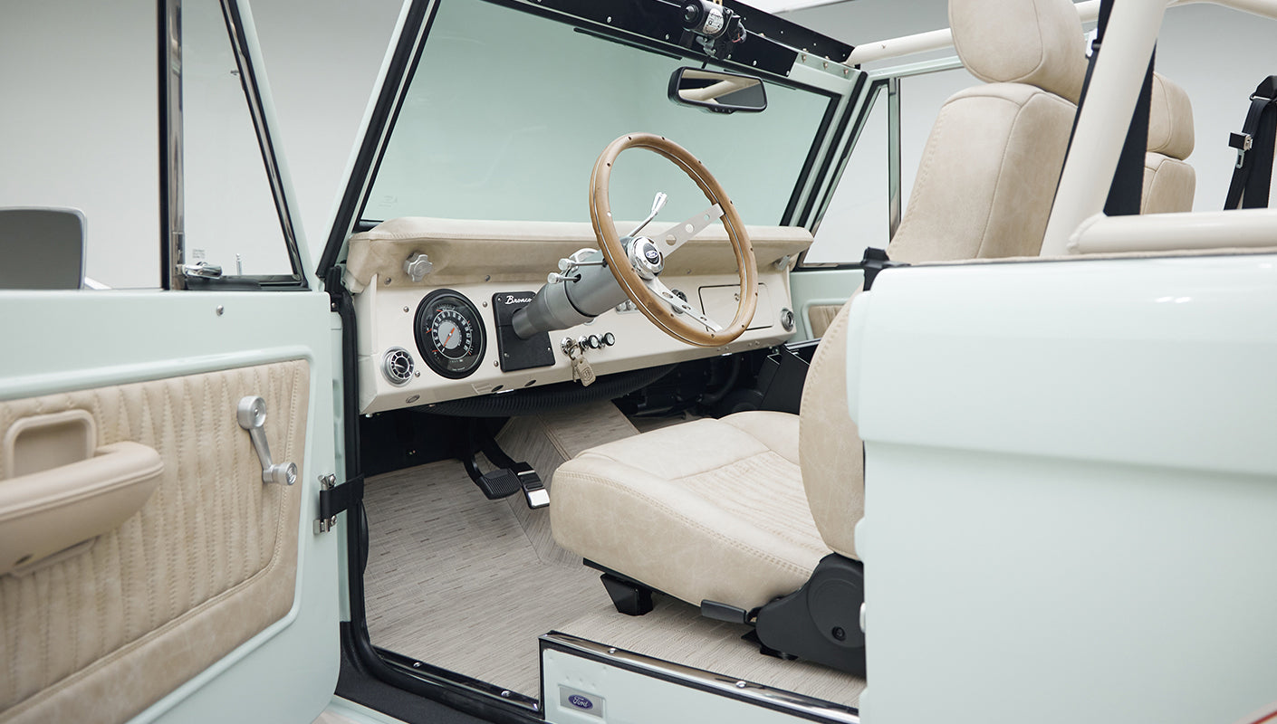 Driver side interior of Classic Ford Bronco with beige seats, wooden steering wheel, and light blue door frame