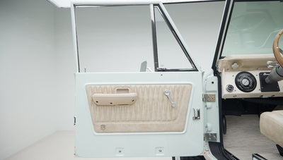 Open driver side door of Classic Ford Bronco showing beige interior panel with stitched pattern and manual window crank