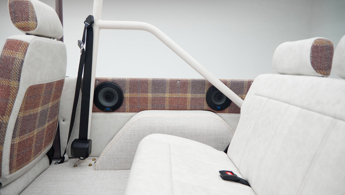 Interior rear passenger area of Classic Ford Bronco with off-white and plaid upholstery, seat belt, and speakers.