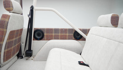 Interior rear passenger area of Classic Ford Bronco with off-white and plaid upholstery, seat belt, and speakers.