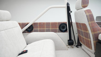 Classic Ford Bronco rear passenger seat and door panel with off-white and plaid fabric, visible seat belt and speakers.