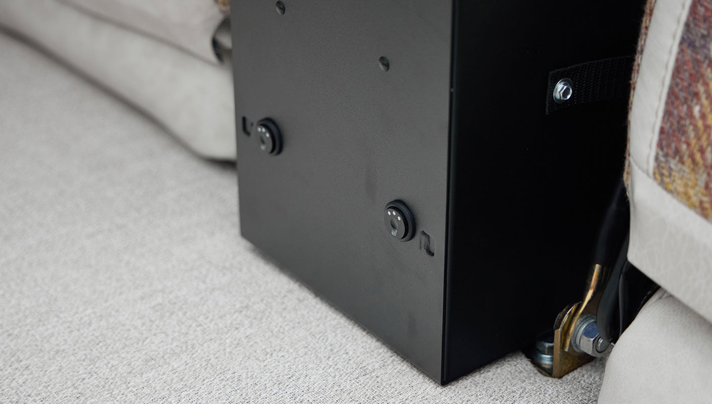Black box under a seat with two round switches having heated seat icons on a textured gray carpeted floor.