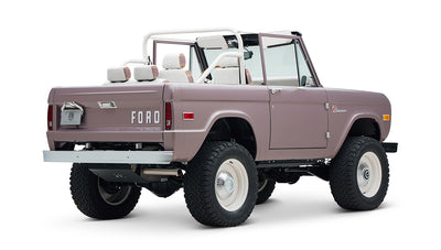 Angled rear of soft pink Classic Ford Bronco showing white wheels, roll bar, and open top on white background