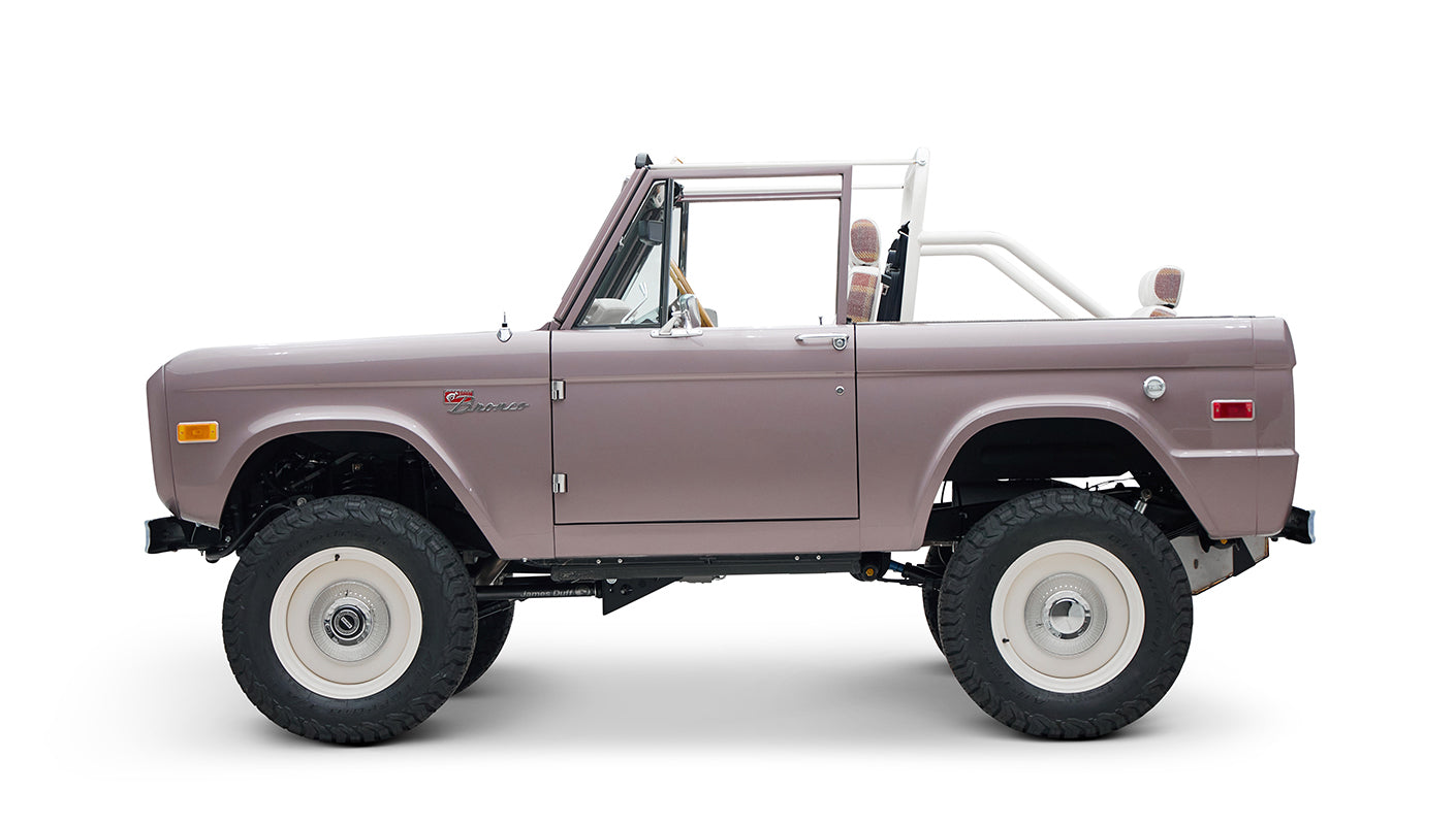 Side view of soft pink Classic Ford Bronco with white wheels, lifted suspension, and no roof on a white background