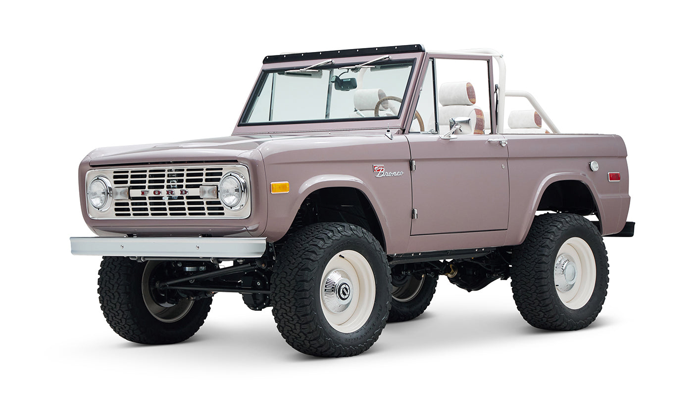 Front view of a soft pink Classic Ford Bronco with round headlights, white wheels, and lifted suspension on white background