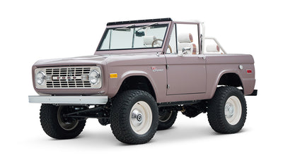Front view of a soft pink Classic Ford Bronco with round headlights, white wheels, and lifted suspension on white background