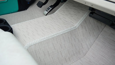 Floor area beneath steering wheel with cream textured floor mat and black pedals in Classic Ford Bronco