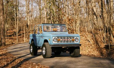 Frozen blue Classic Ford Bronco on paved road with leafless trees in background