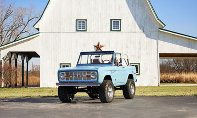 Frozen blue Classic Ford Bronco parked on empty pavement in front of white barn with green window frames