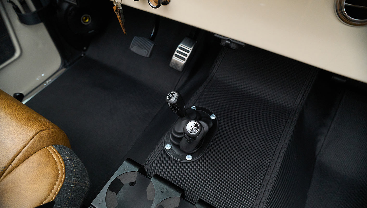 Floor area showing two gear shifters mounted on black mat with tan and plaid seat edges visible