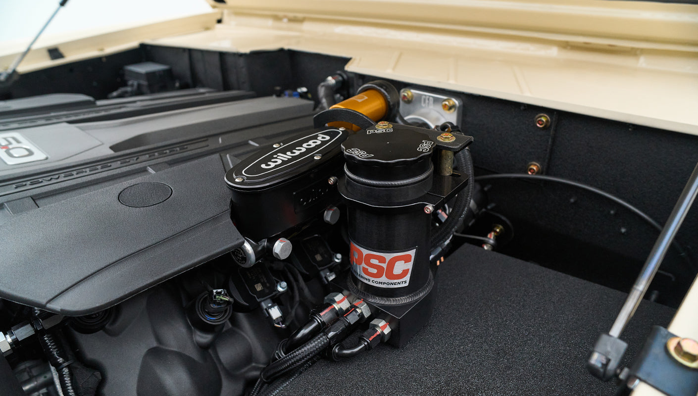 Close-up of car engine components including a black Wilwood part and a PSC reservoir