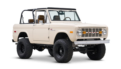Cream Classic Ford Bronco with black roll bars, off-road tires, and tan seats, angled front side view