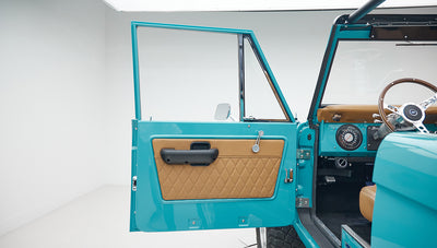 Open driver side door of Classic Ford Bronco showing tan door panel and peacock blue exterior.