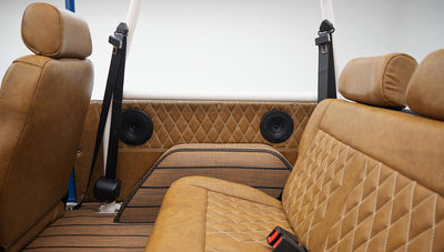 Rear interior view showing tan leather seats, woven floor mats, seat belts, and white roll cage in Classic Ford Bronco