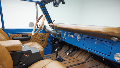 Front interior of Classic Ford Bronco with tan leather seats, wood steering wheel, and blue dashboard