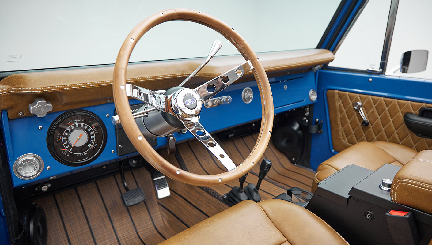 Classic Ford Bronco steering wheel with wood finish and chrome spokes, blue dashboard, and tan leather seats