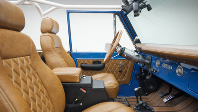 Front seats and blue dashboard of Classic Ford Bronco showing wood-look steering wheel and tan leather