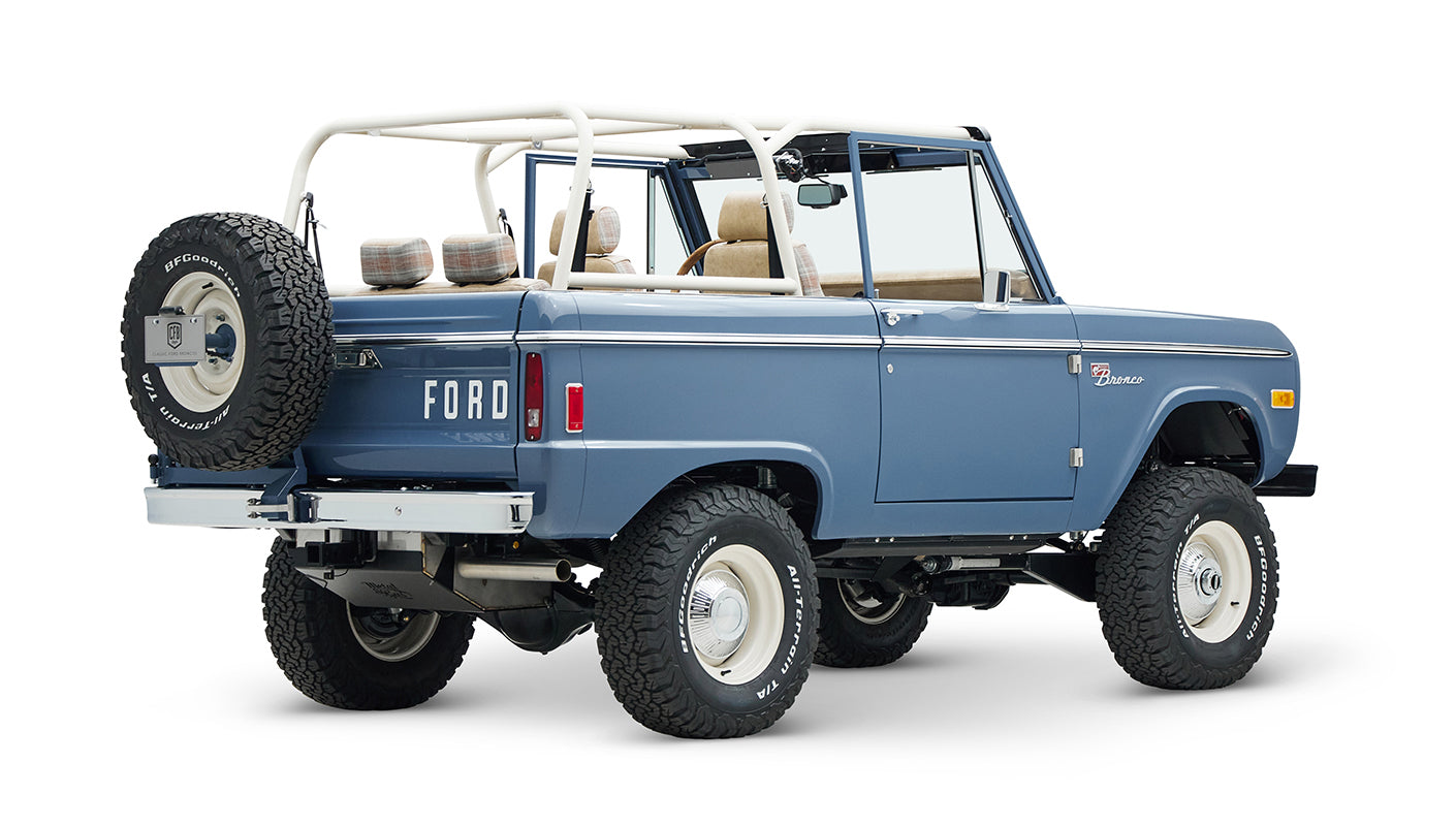 Classic Ford Bronco in light blue, rear side view showing white roll cage and spare tire with large off-road tires.