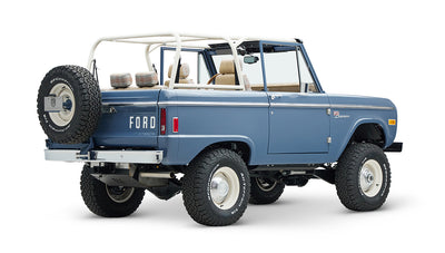 Classic Ford Bronco in light blue, rear side view showing white roll cage and spare tire with large off-road tires.