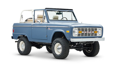Classic Ford Bronco in light blue, front side angle with white roll cage and tan plaid seats.