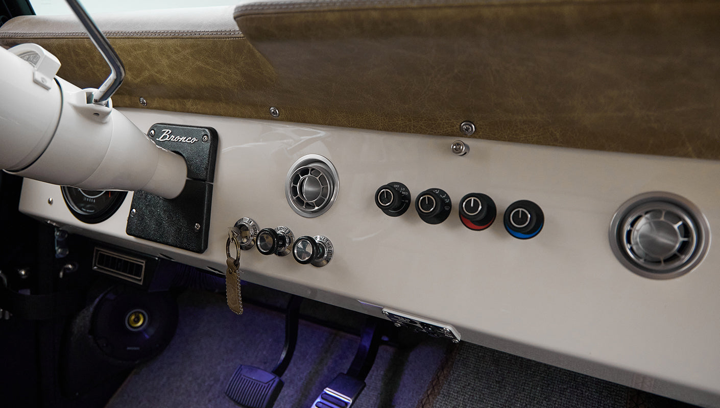 Dashboard controls, air vents, and ignition key switch inside Classic Ford Bronco with tan and white interior