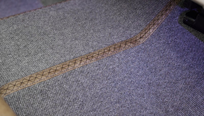 Close-up view of a textured gray automotive carpet with stitched brown edges