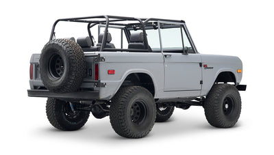 Rear three-quarter view of a Classic Ford Bronco in gray with black wheels, roll cage, and spare tire mounted on back